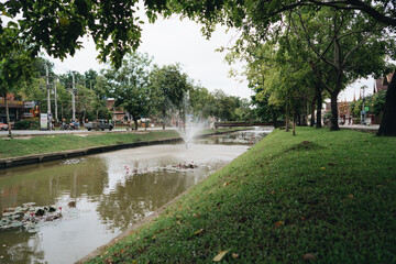 Fototapeta na wymiar View on a channel with fountains, big trees and streets on the banks. Chiang Mai