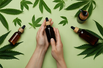Female hands hold bottle with cannabis oil on green background with leaves and bottles