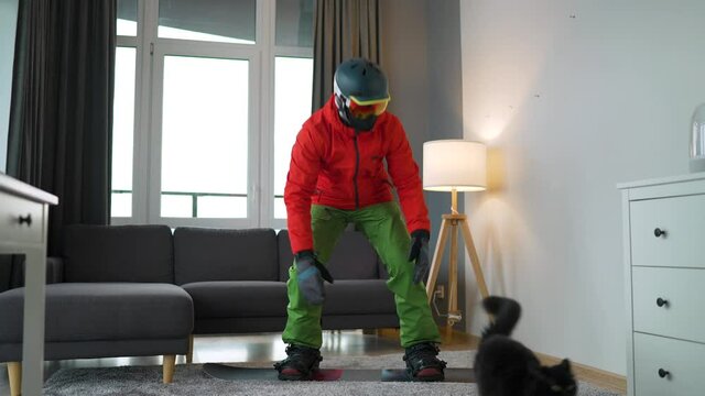 Funny video. Man dressed as a snowboarder depicts snowboarding on a carpet in a cozy room. He plays with a black fluffy cat. Waiting for a snowy winter. Slow motion