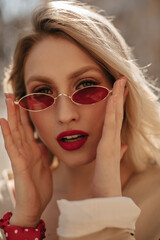 Grey-eyed blonde curly woman with red lips looks sensitively into camera and puts on bright colorful sunglasses outside.