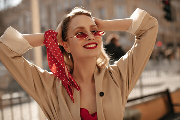 Good-humored blonde woman in beige trench coat, silk blouse and bright sunglasses smiles sincerely...