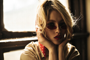 Young serious blonde woman in beige trench coat and red sunglasses looks into camera. Curly lady...