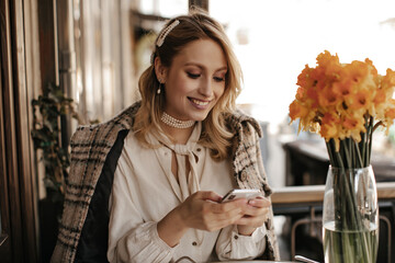 Cheerful blonde woman in tweed checkered coat, white blouse and pearl necklace sits in cozy cafe...