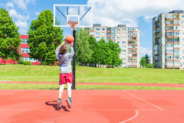 Cheerful Boy performs shot to the basket at basketball game on the playground during sunny summer...