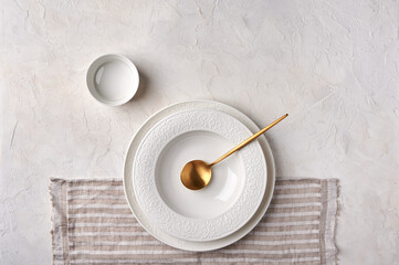Empty white plate, gravy boat, golden spoon on napkin. Kitchen utensils set on light gray table. Top view flat lay with copy space