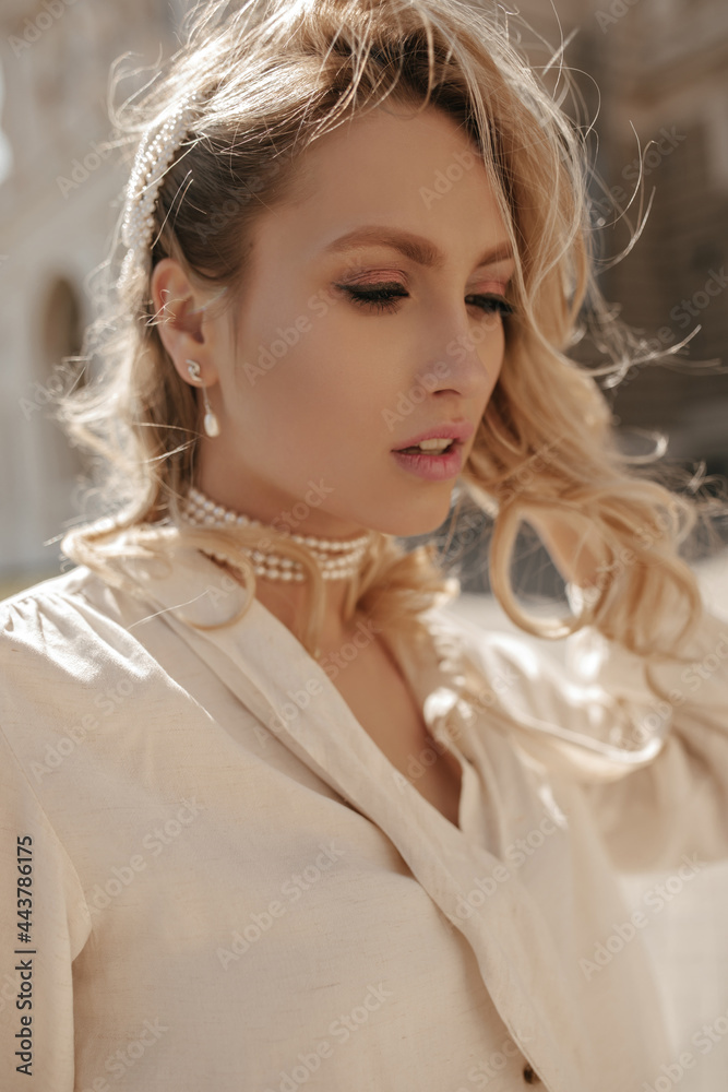 Wall mural Attractive pretty blonde woman in white blouse and pearl necklace looks down and walls outside.