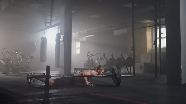 A young man without a shirt in slow motion jumps a burpee over a barbell in the gym in backlight