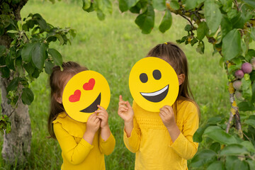 Two children in the park are standing with their faces covered with happy emoticons and showing hand gestures.  Funny temperamental girls. Communication. 