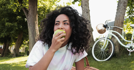 young african american woman eating fresh apple near bicycle in park.