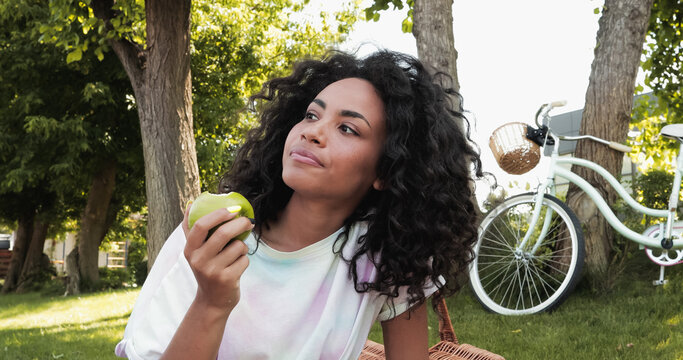 Curly African American Woman Eating Fresh Apple Near Bicycle In Park.