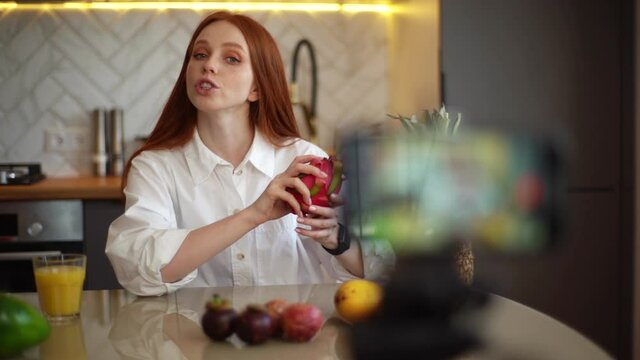 Close-up of female vlogger shooting culinary show speaking about exotic fruits looking at camera. Redhead young woman blogger recording live tutorial video about healthy vegetarian eating.