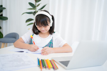 Little asian girl smiling and writing a book. She is studying online at home.