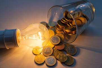 Incandescent light bulb turned on, with jar full of coins next to it, on wooden surface. Increased costs. Savings. Energy tariffs.