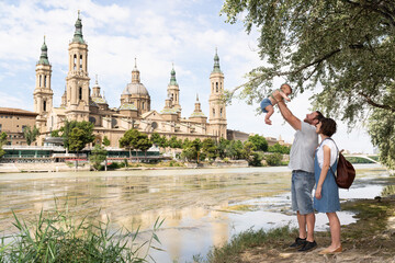 Young family playing with their baby at the Ebro river bank with Basilica del Pilar on the background. Family vacation in Zaragoza, Spain.