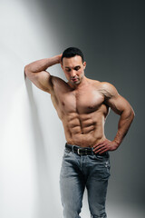 A young athlete bodybuilder poses in the studio topless in jeans near the wall. Sport.