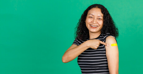 Asian middle aged woman smiling and pointing at her arm with bandage patch showing she got vaccinated for Covid 19 virus on green background. Concept for Covid 19 vaccination