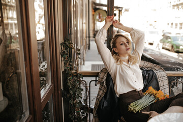 Attractive curly blonde lady in pearl necklace and white blouse rises arms and sits in cozy street cafe with yellow bouquet.