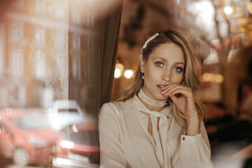 Blonde grey-eyed charming lady in vintage white blouse and pearl necklace gently looks into camera inside.