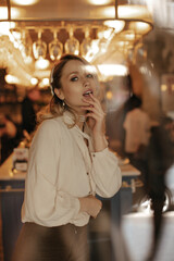 Beautiful blonde woman in brown velvet pants and white blouse looking into camera and gently touches lips. Attractive lady in pearl necklace poses in restaurant.