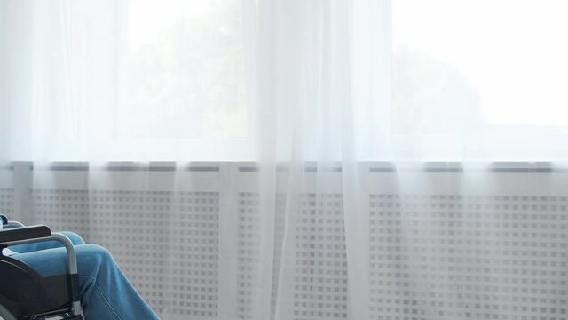 Disabled old man is sitting in a wheelchair at home alone. A handicapped person in a wheelchair is looking out the window. Paralysis and disability.