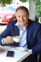 A young man in a blue business suit with a cup of coffee sitting in a summer cafe. Coffee break. Businessman. Morning business people concept