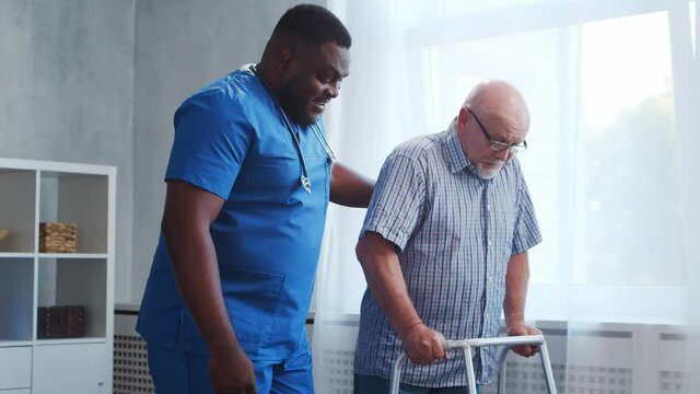 African-American caregiver is teaching disabled old man to walk with walker. Professional nurse and handicapped patient in a nursing home. Assistance, rehabilitation and health care.