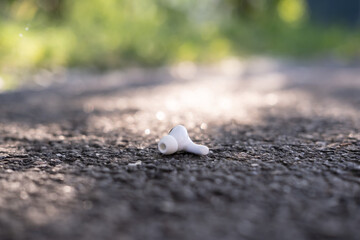 One wireless headphones is lost and lies on the asphalt sidewalk, outdoors.