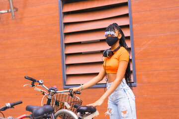 Fototapeta na wymiar Black breed girl with mask by coronavirus pandemic, african ethnic group with orange shirt in the city. Cofing the bike parked in the city