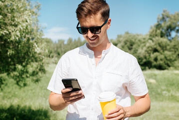 Portrait of happy young man browsing Internet on mobile phone and holding disposable cup of coffee in nature, outdoors. Smiling man with glasses using smartphone on vacation in the park on summer day