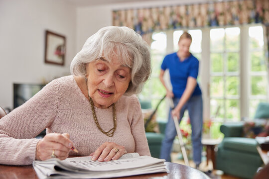 Female Home Help Cleaning House With Vacuum Cleaner Whilst Senior Woman Does Crossword In Newspaper