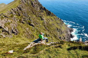 Obraz premium Bald male tourist in green shirt sitting on the edge of a rock. Achill island, county Mayo, Ireland. Warm sunny day, blue sky and water. Man reckless behavior concpet