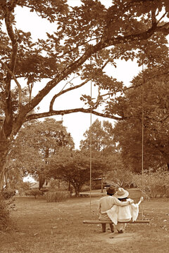 Sepia Image Of Happy Couple Sitting Side By Side On A Wooden Swing In The Garden