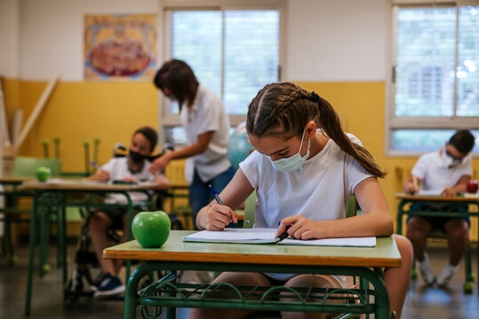 A girl student, wearing a protective mask, sits at a desk writing in her notebook in the background a teacher helps a disabled student sitting in a wheelchair. Back to school concept 