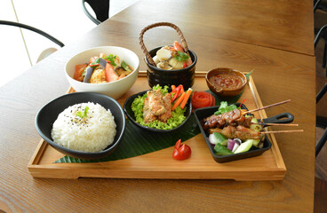 asian bento party set with seafood soup, deep fried snack, chicken satay and peanut spicy sauce, stir fried vegetables and steam white rice on wood board tray asian halal menu