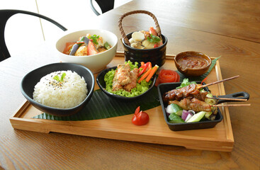 asian bento party set with seafood soup, deep fried snack, chicken satay and peanut spicy sauce, stir fried vegetables and steam white rice on wood board tray asian halal menu
