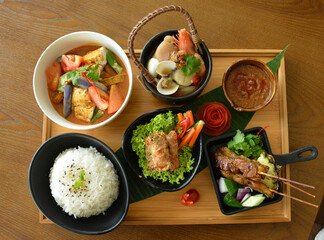 asian bento party set with seafood soup, deep fried snack, chicken satay and peanut spicy sauce, stir fried vegetables and steam white rice on wood board tray asian halal menu