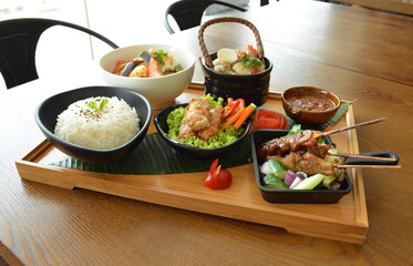 asian bento party set with seafood soup, deep fried snack, chicken satay and peanut spicy sauce, stir fried vegetables and steam white rice on wood board tray asian halal menu
