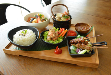 asian bento party set with seafood soup, deep fried snack, chicken satay and peanut spicy sauce, stir fried vegetables and steam white rice on wood board tray asian halal menu