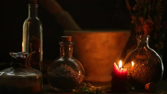 Bottles filled with herbs and a mortar and pedestal illuminated by a single red candle on an alchemy worktable