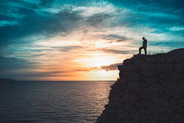 Man silhouette stands on the edge of the abyss and looks the sea with beautiful colorful sky.