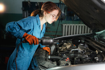 A girl auto mechanic in a repair shop or garage inspects the engine of a car, illuminating with a special lamp. Locksmith woman at the workplace works. Maintenance and service of cars.