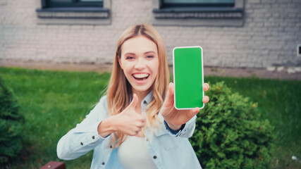 Fototapeta na wymiar blurred and cheerful student holding smartphone with green screen while showing thumb up near building
