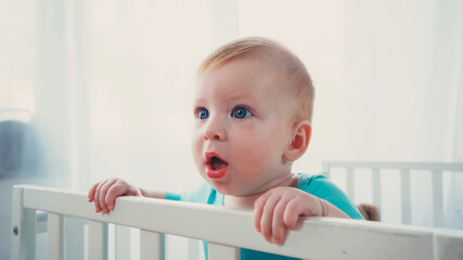 surprised infant boy standing in baby crib and looking away - obrazy, fototapety, plakaty