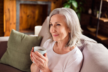 Portrait of attractive dreamy grey-haired woman sitting on divan drinking cacao nice day at home...