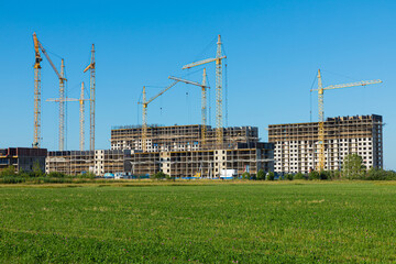 Cloudless landscape or scene of meadow with green grass and bushes with concrete modern building construction with tall yellow cranes on the background of clear blue sky