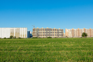 Cloudless urban landscape or scene of meadow with green grass, bushes, alone tree and crane with concrete modern buildings on the background of clear blue sky. New block or district in the edge of the