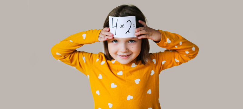 funny crazy cute kid girl in mustard dress learns the multiplication table on beige background, child with an example sheet and numbers in his hands