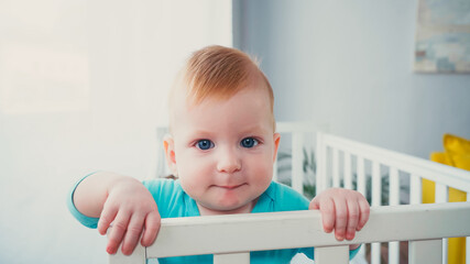 joyful infant boy standing in baby crib and looking at camera - obrazy, fototapety, plakaty