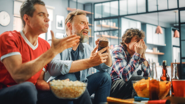 Home: Three Soccer Fans Sitting on a Couch Watch Game on TV, Use Smartphone App to Online Bet, Worrying for Their Soccer Team. Friends Cheer, Shout, Watch Football Play.