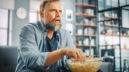 Handsome Middle Aged Bearded Man Sitting on a Couch Watches TV at Home. Focused Fan Eats Snacks,...